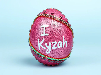 Pink glitter egg encrusted with pink ruby garnets, with spiral bands of garnetts wrapped around in a helix with colors arranged as rainbow stripes, with the words "I ♥️ Kyzah" painted on the egg