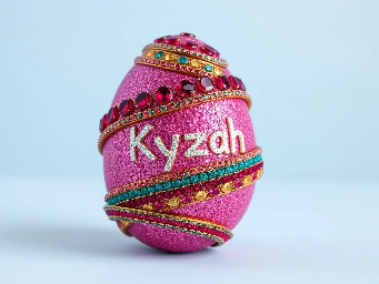 Pink glitter egg encrusted with pink ruby garnets, with spiral bands of garnetts wrapped around in a helix with colors arranged as rainbow stripes, with the name Kyzah painted on the egg